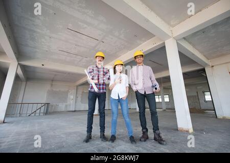 Équipe d'ingénieurs de la diversité, une femme et deux hommes, debout ensemble et souriant à la caméra tout en travaillant sur le chantier de construction d'un bâtiment intérieur. Banque D'Images