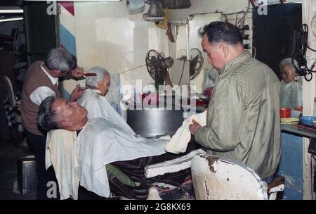 1987, historique, Hong Kong, à l'intérieur d'un ancien coiffeur traditionnel, un homme asiatique âgé ayant une coupe de cheveux et un autre homme asiatique, assis sur une chaise pour un rasage sur peau humide. Il est à noter que la chaise Barbie ancienne avec dossier inclinable est importante pour les clients qui se rasent, car la chaise inclinable est plus sûre. Banque D'Images