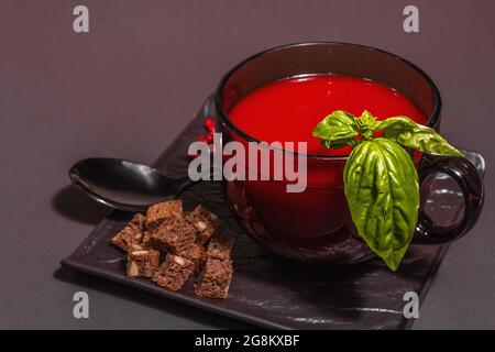 Soupe traditionnelle de tomates dans une tasse. Basilic, croûtons, épices, couverts. Fond en béton de pierre noire, design minimaliste, espace de copie Banque D'Images