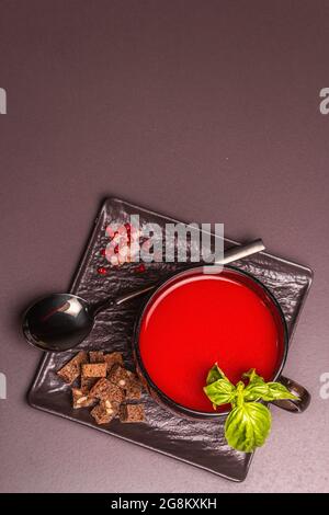 Soupe traditionnelle de tomates dans une tasse. Basilic, croûtons, épices, couverts. Fond en béton de pierre noire, design minimaliste, vue de dessus Banque D'Images