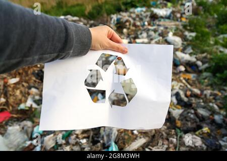 Main tenant le papier coupé avec le logo de recyclage sur l'arrière-plan de la poubelle de vidage. Concept de recyclage. Pollution de la planète. Banque D'Images