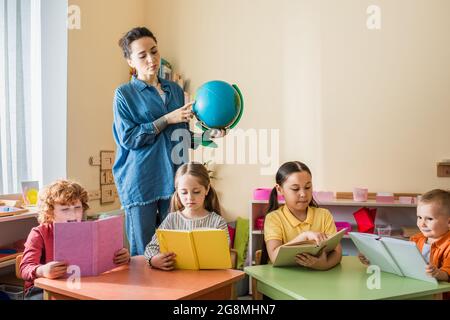 professeur pointant vers le monde près des enfants multiethniques lisant des livres pendant la leçon Banque D'Images