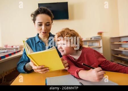 redhead garçon tenant un stylo et un livre de lecture entre les mains d'un jeune enseignant Banque D'Images