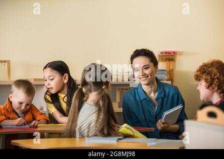 jeune professeur avec livre souriant près des enfants interraciaux dans la salle de classe de montessori Banque D'Images