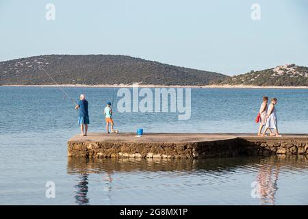 Vodice, Croatie - 8 juillet 2021 : homme âgé et jeune garçon pêchant sur une jetée tandis que les femmes âgées marchent Banque D'Images