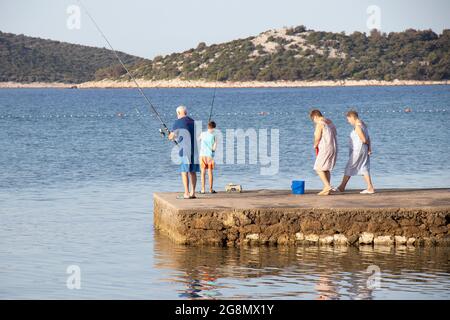 Vodice, Croatie - 8 juillet 2021 : homme âgé et jeune garçon pêchant sur une jetée tandis que les femmes âgées marchent Banque D'Images