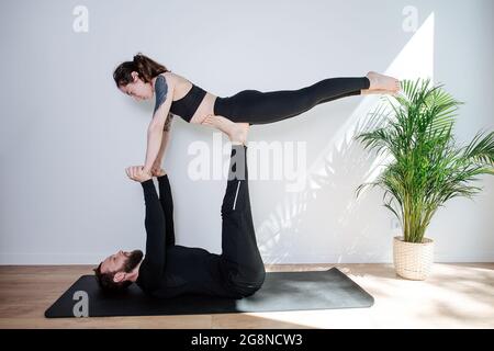 Homme et femme faisant la forme de gymnastique, il la tient sur les bras et les jambes de strtaight. Dans un vêtement de sport noir. Devant un mur blanc. Banque D'Images