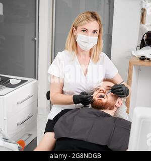 Femme stomatologiste dans le masque médical regardant la caméra tout en examinant les dents de patient mâle avec l'explorateur dentaire et le miroir. Concept de dentisterie, stomat Banque D'Images