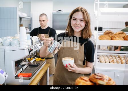 Jeune belle boulangère fille posant avec le café et la carafe de crème dans les mains. Charmant sourire à l'appareil photo. Dans une cuisine de boulangerie désordonnée. Banque D'Images