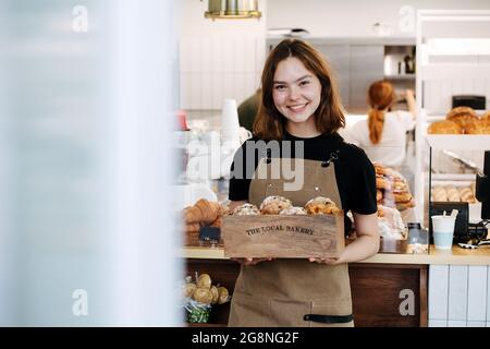 Petite fille sympathique posant avec une boîte en bois de marque, remplie de muffins. Elle porte un tablier et regarde la caméra. Dans une boulangerie très fréquentée. Banque D'Images