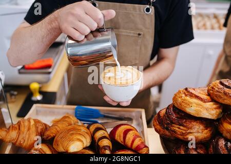 Image rognée d'un boulanger masculin mettant de la crème dans une tasse de café d'une verseuse métallique. Dans une cuisine de boulangerie à côté d'une pile de pâtisserie. Banque D'Images