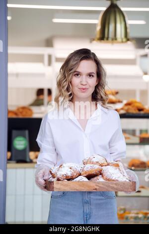 Portrait de bokeh d'une femme blonde posant avec un plateau rempli de pâte à un seuil de son magasin. Elle porte une chemise habillée et un Jean léger, regardant le Banque D'Images