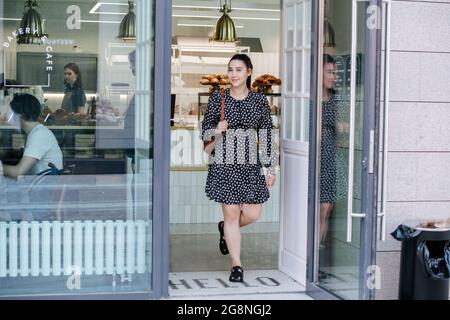 Femme satisfaite, sortant du caffee. Elle porte une robe d'été à motif fleuri. Tenue d'un manteau dans ses mains Banque D'Images