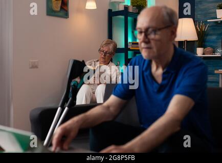Personnes âgées en retraite dans la salle de séjour à la maison. Femme caucasienne en fauteuil roulant de lecture et homme vieux regardant le périphérique virtuel ordinateur portable Internet avec des béquilles Banque D'Images