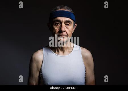 Athlète âgé portant une chemise sans manches blanche, avec des marques de soleil sur les bras et un bandeau bleu, sur un fond sombre. Sport et victoire concept. Banque D'Images