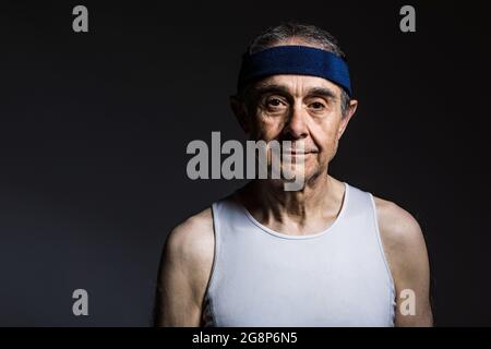 Athlète âgé portant une chemise sans manches blanche, avec des marques de soleil sur les bras et un bandeau bleu, sur un fond sombre. Sport et victoire concept. Banque D'Images