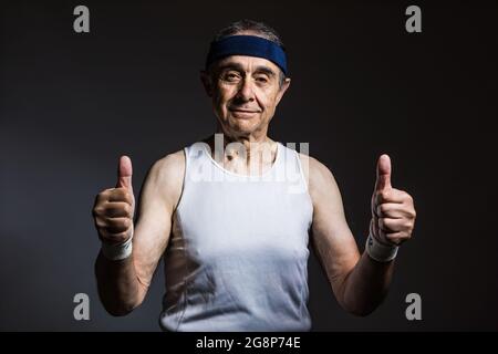 Athlète âgé portant un débardeur blanc, avec des marques de soleil sur les bras, et un bandeau bleu, pouces vers le haut, sur un fond sombre. Sport et victoire concept. Banque D'Images