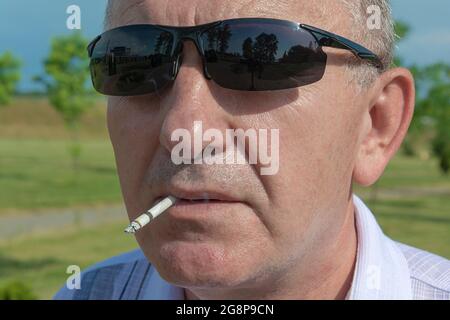 l'homme tient une cigarette dans la bouche. Le visage d'un homme en lunettes de soleil est gros plan sur un arrière-plan flou. Photo de stock Banque D'Images