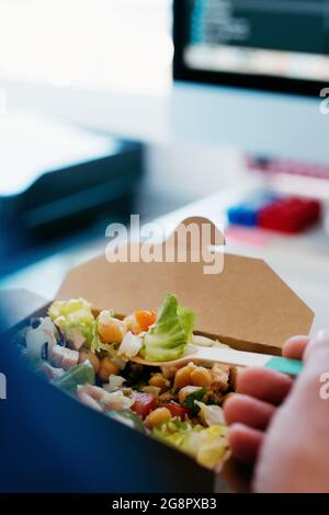 gros plan d'un jeune homme du caucase mangeant une salade de pois chiches dans un contenant en papier, à l'aide d'une fourchette en bambou, assis à son bureau Banque D'Images