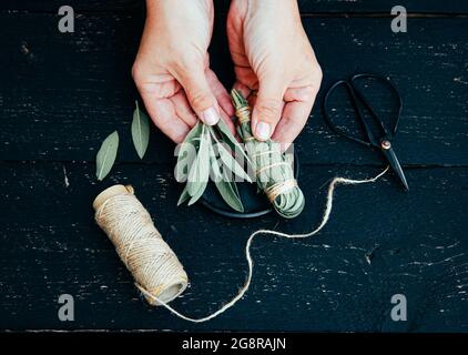 Personne faisant la sauge blanche Salvia apiana bâton à la maison avec des feuilles de sauge maison. Ficelle de coton et ciseaux vintage pour la décoration sur le noir. Banque D'Images