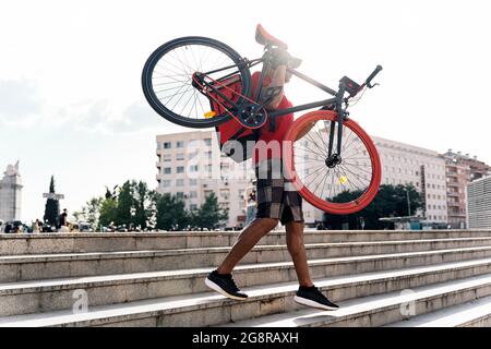 Un messager non reconnu portant une casquette portant son vélo dans la ville pour livrer un paquet. Banque D'Images