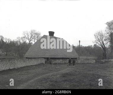 Années 1950, historique, deux dames pour une promenade sur un chemin herbeux sur les bas nord près de Nore, Kent, en passant devant un mur de pierre à côté d'un cottage ou d'un vieux bâtiment de ferme avec un toit de chaume inhabituel et grand. Banque D'Images