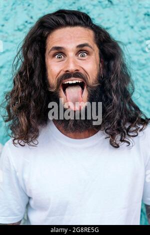 Homme barbu frais avec de longs cheveux qui collent sa langue et regardant l'appareil photo contre le mur bleu. Banque D'Images