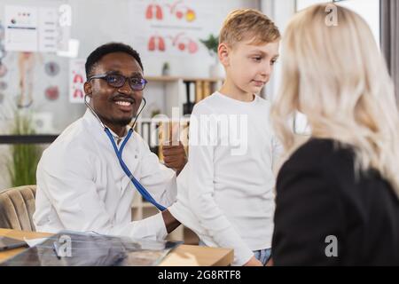 Concept de soins de santé et de pédiatrie. Souriant confiant, le médecin généraliste africain vérifie les poumons et la respiration du jeune garçon lors de sa visite régulière prévue à la clinique familiale avec sa mère Banque D'Images