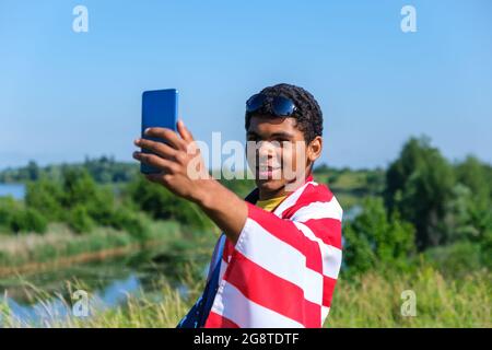 Jeune homme afro-américain souriant couvert d'un drapeau américain prenant le selfie Banque D'Images