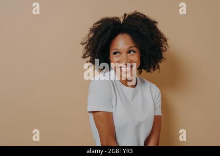 Mignonne africaine avec un sourire crasseux regardant de côté avec de la timidité, isolée sur fond beige Banque D'Images