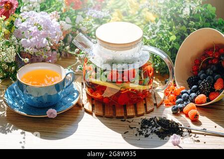 thé aux fruits aromatiques naturels dans une théière transparente et dans une tasse parmi les baies et les fleurs. thé aromatique réchauffant avec un arôme profond de baies et de fleurs sauvages Banque D'Images