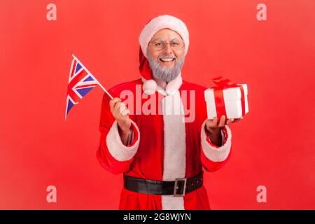 Patriotique heureux homme âgé avec barbe grise portant le costume du père noël tenant le drapeau de la Grande-Bretagne et enveloppé boîte cadeau, cadeau de Noël. Indo Banque D'Images