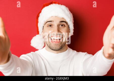 Gros plan d'un beau homme dans un chapeau de père noël, prenant un selfie ou ayant un appel vidéo, tenant l'appareil photo des deux mains, debout sur fond rouge Banque D'Images