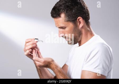 Homme dans un T-shirt blanc utilisant un compte-gouttes d'huile facial pour faire tomber le liquide dans ses mains. Banque D'Images