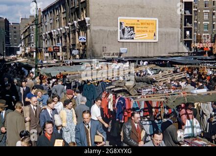 Les amateurs de shopping sont à la recherche d'un marché très fréquenté de Petticoat Lane, un marché de rue général et de vêtements à Spitalfields, dans l'est de Londres, Angleterre, Royaume-Uni vers 1955. Il se compose de deux marchés de rue adjacents. Wentworth Street Market et Middlesex Street Market. Le dimanche, il s'étend sur de nombreuses rues environnantes, avec plus d'un millier d'étals. En arrière-plan se trouve un palissade ou un panneau publicitaire annonçant la bière brune de Watney. Cette image est tirée d'une ancienne transparence couleur Kodak 35 mm amateur, une photographie vintage des années 1950. Banque D'Images