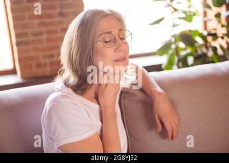 Portrait d'une femme malade aux cheveux gris, assise sur un divan touchant le cou se sentant mal à la maison à l'intérieur Banque D'Images