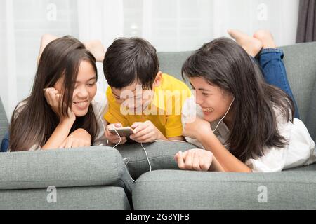 Trois enfants de la famille asiatique, un garçon et deux filles jouent avec le bonheur à la maison, en utilisant le téléphone intelligent pour écouter de la musique et regarder des vidéos Banque D'Images