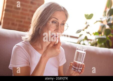 Portrait de la femme attrayante en bonne santé à cheveux gris assis sur divan prenant des pilules médicam à loft maison industrielle à l'intérieur Banque D'Images