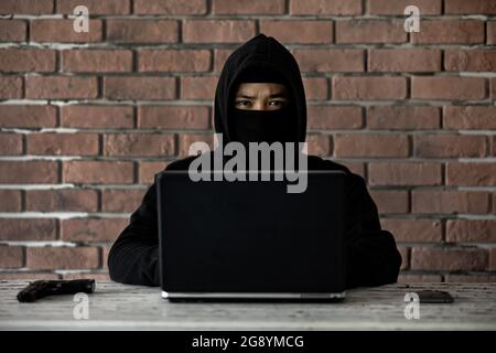 Cyber-voleur dans la capuche noire avec ordinateur portable, pistolet et téléphone mobile regarder à la caméra. Concept de crimes sur Internet et de manière illégale. Banque D'Images