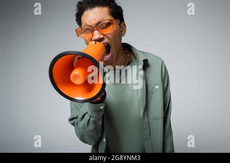 jeune afro-américain en lunettes stylées criant en mégaphone isolé sur gris Banque D'Images