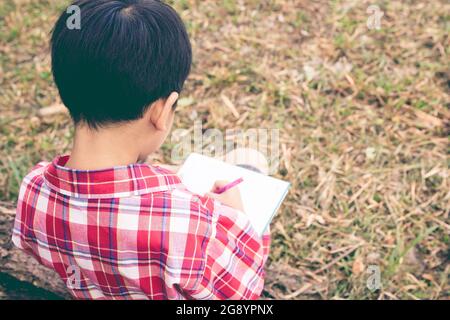 Vue arrière. Enfant utilisez un stylo pour écrire sur un carnet en bois dans le parc. À l'extérieur, en journée, en plein soleil. Les enfants lisent et étudient, educati Banque D'Images