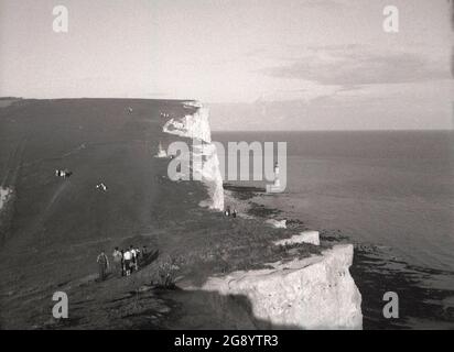 Années 1950, photo historique , les gens à pied à Beachy Head, près d'Eastbourne, East Sussex, Angleterre, Royaume-Uni. La plus haute falaise de la mer de craie en Grande-Bretagne, la tournière ondulée est un endroit célèbre pour la promenade de plaisir, avec une vue magnifique sur la Manche. Une zone d'une beauté naturelle exceptionnelle, en 1929, une loi du Parlement, a permis l'achat des terres pour protéger et sauvegarder leur utilisation pour les générations futures. Le phare offshore construit en 1902 peut être vu au loin. Banque D'Images