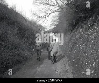 Années 1950, historique, à l'extérieur dans une coupe dans la campagne, près de Nore, Kent, Englod, deux hommes, l'une avec un sac à dos en toile et l'autre portant un long manteau et avec une sacoche, et une dame marchant le long d'un chemin ou d'une piste abritée, peut-être autrefois une petite rivière qui a séché. Devant eux, un pont couvert encadré en bois traversant le chemin. Banque D'Images