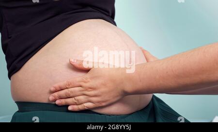 Le médecin vérifie que la femme enceinte touche un grand ventre par les mains assises près du mur vert clair dans le bureau de la clinique à la clôture du rendez-vous Banque D'Images