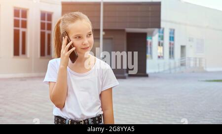 Une fille parle sur son smartphone et sourit. Blonde écofille avec de longs cheveux en queue de cheval parle avec un ami via le microphone gadget debout dans la rue, copyspace Banque D'Images