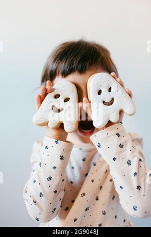 Petite fille avec la bouche ouverte couvrant les yeux avec des biscuits fantômes Banque D'Images