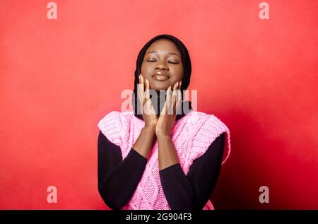 Jeune femme avec les yeux fermés touchant les joues sur fond rouge Banque D'Images
