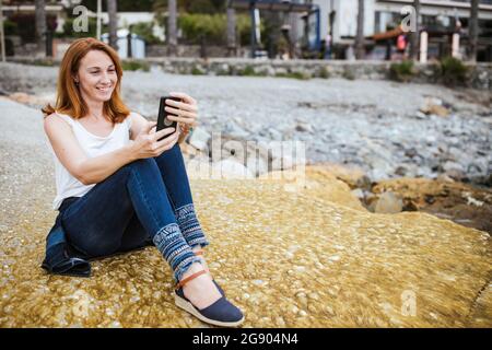 Femme souriante prenant le selfie à travers le smartphone tout en étant assise sur la voie des pieds Banque D'Images