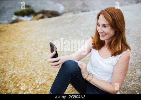 Une femme souriante qui prend son selfie à travers un téléphone portable tout en étant assise sur un sentier Banque D'Images