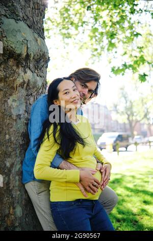 Femme enceinte souriant pendant que son mari se serre de derrière dans le parc public Banque D'Images